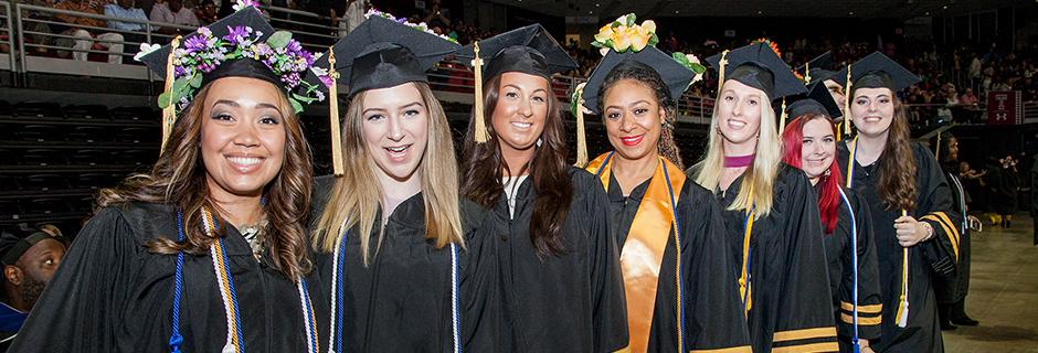 A row of female students wearing caps and gowns at graduation, all sanding up and smiling at the camera.
